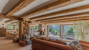 A rustic interior with exposed wooden beams, a stone wall, a fireplace, and large windows offering a mountain view.