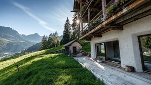 A wooden chalet with a balcony adorned with flowers, a stone cottage, and a view of mountains.