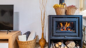 A modern living room with a black wood-burning stove, a flat-screen TV, a wicker basket with cushions, and a vase with branches. - Casa en las Montañas de Benicassim — Mountain Forest Retreat with Private Terrace and Wi-Fi (Benicassim)