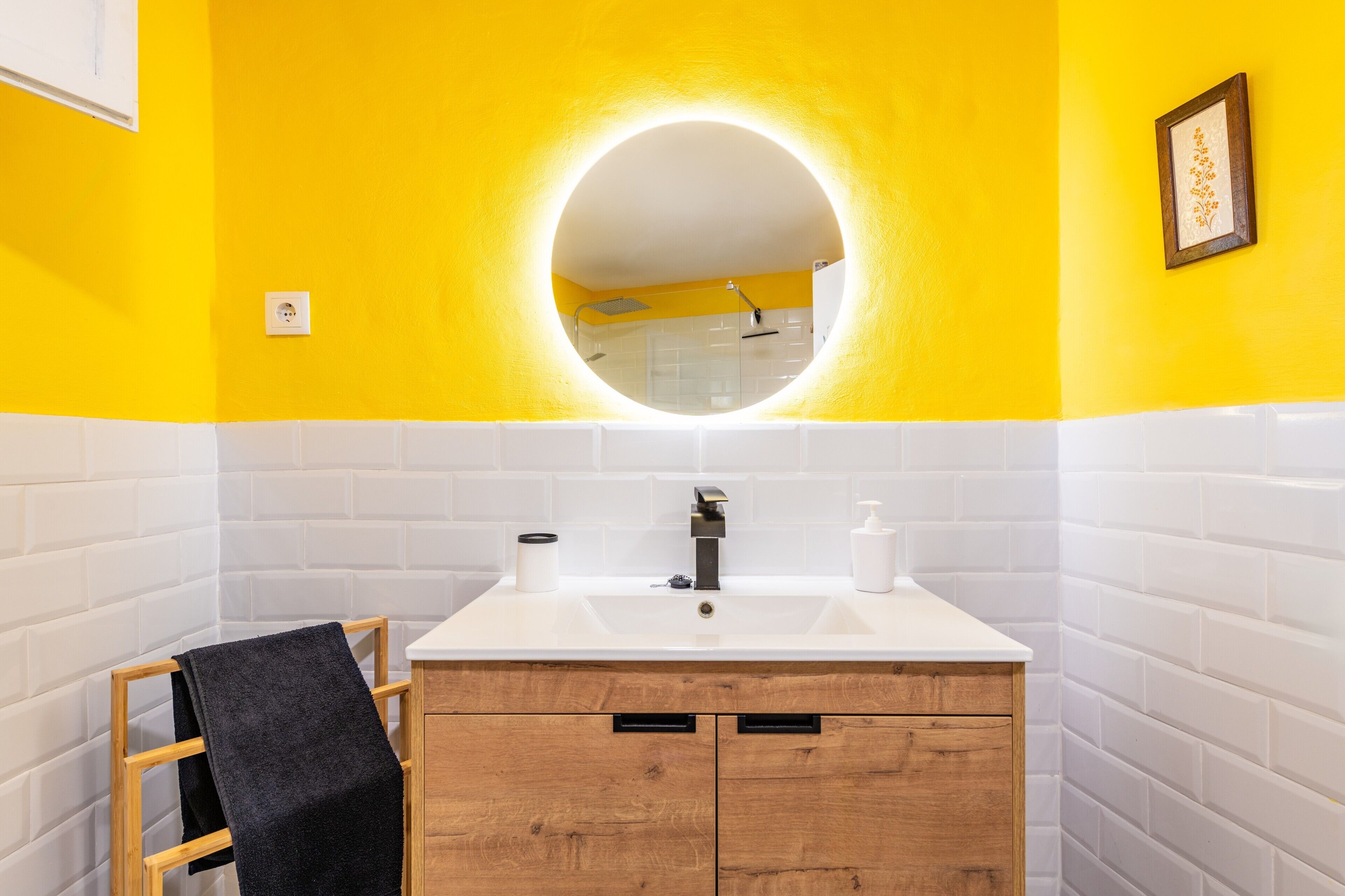 Een badkamer met een witte wastafel, houten kastjes, een zwarte handdoek en een ronde spiegel met verlichting.
