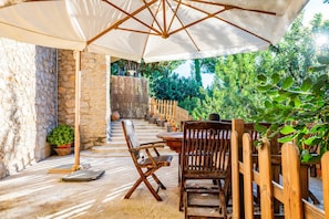 Une terrasse extérieure avec un mur en pierre, des chaises en bois, une table et un grand parasol.