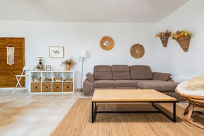 Un salon avec un canapé marron, une table basse en bois, une table d’appoint blanche avec des paniers, un meuble blanc et des œuvres d’art décoratives au mur.