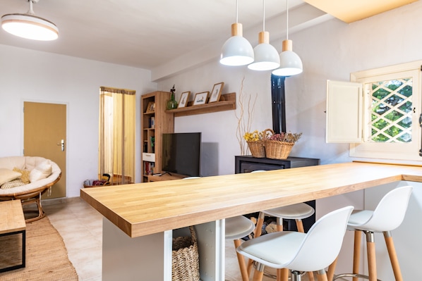 Une cuisine moderne avec un plan de travail en bois, des chaises blanches, des luminaires suspendus et un téléviseur.