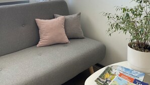 Ein Wohnzimmer mit grauem Sofa, einem rosafarbenen Kissen, einer weißen Wand mit einem Gemälde, einem Fenster mit weißen Vorhängen und einer Zimmerpflanze.