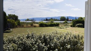 Une vue depuis une fenêtre sur un jardin avec une balançoire, divers arbustes et une chaîne de montagnes au loin.