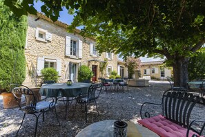 An outdoor patio area with stone buildings, outdoor seating, and a large tree providing shade. - Chambre d'hôtes -B&B - Le Cocon de Manon (Lagnes)