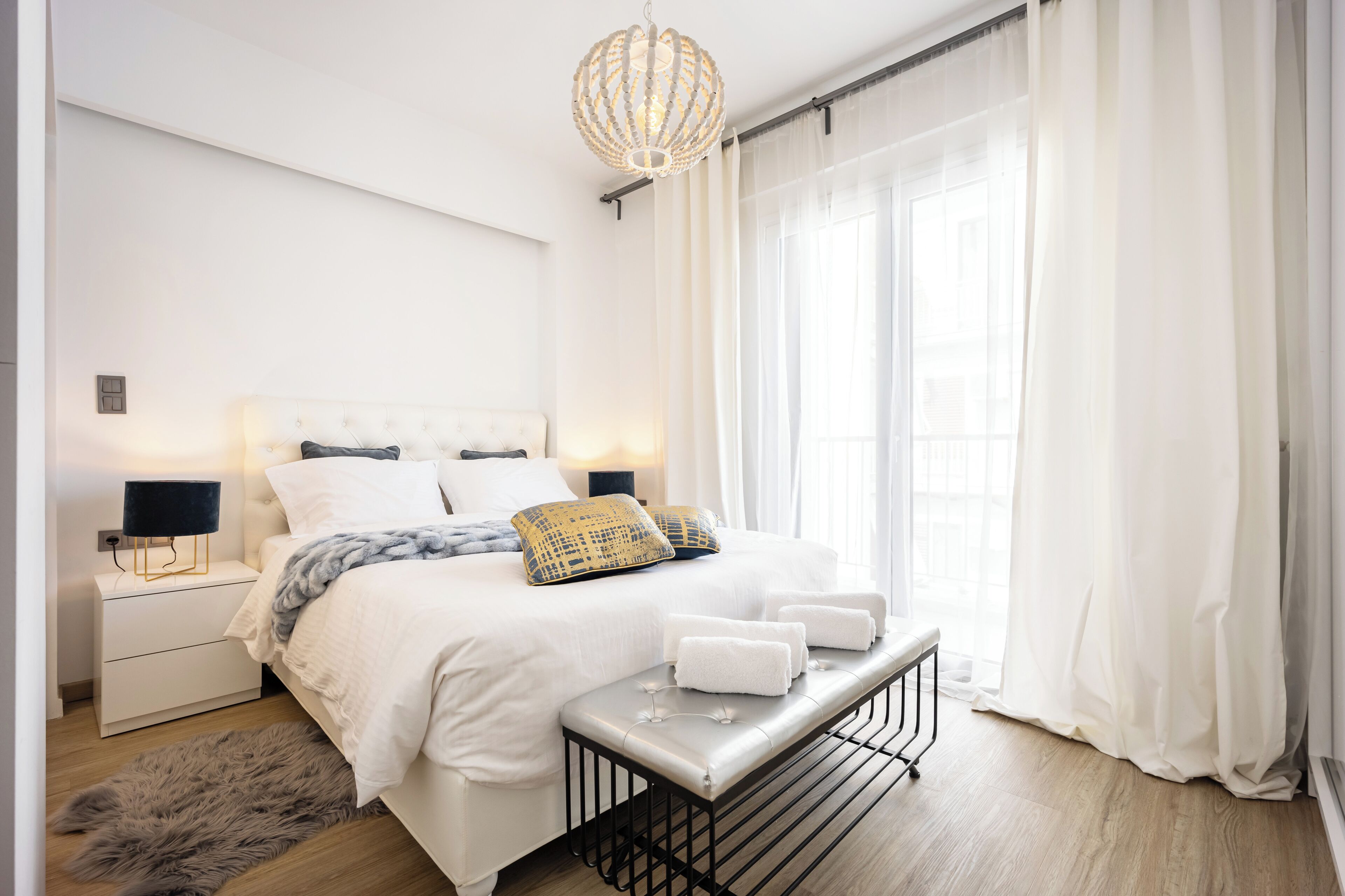 A modern bedroom with a large bed, bedside lamps, a bench with towels, and sheer curtains.