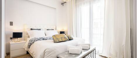 A modern bedroom with a large bed, bedside lamps, a bench with towels, and sheer curtains.