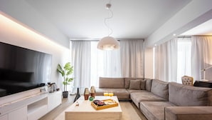 A modern living room with a large flat-screen TV, a sectional sofa, a coffee table with snacks, and a pendant light. - Luxury 3-Bedroom Apartment Athens Nea Smirni (ΝΕΑ ΣΜΥΡΝΗ)