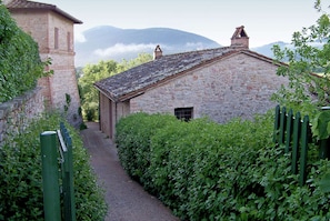 A narrow stone pathway leading to a rustic stone building with a sloped roof, surrounded by lush greenery and a mountain backdrop. - Countryside Retreat With Pool, Mantignana (Corciano)