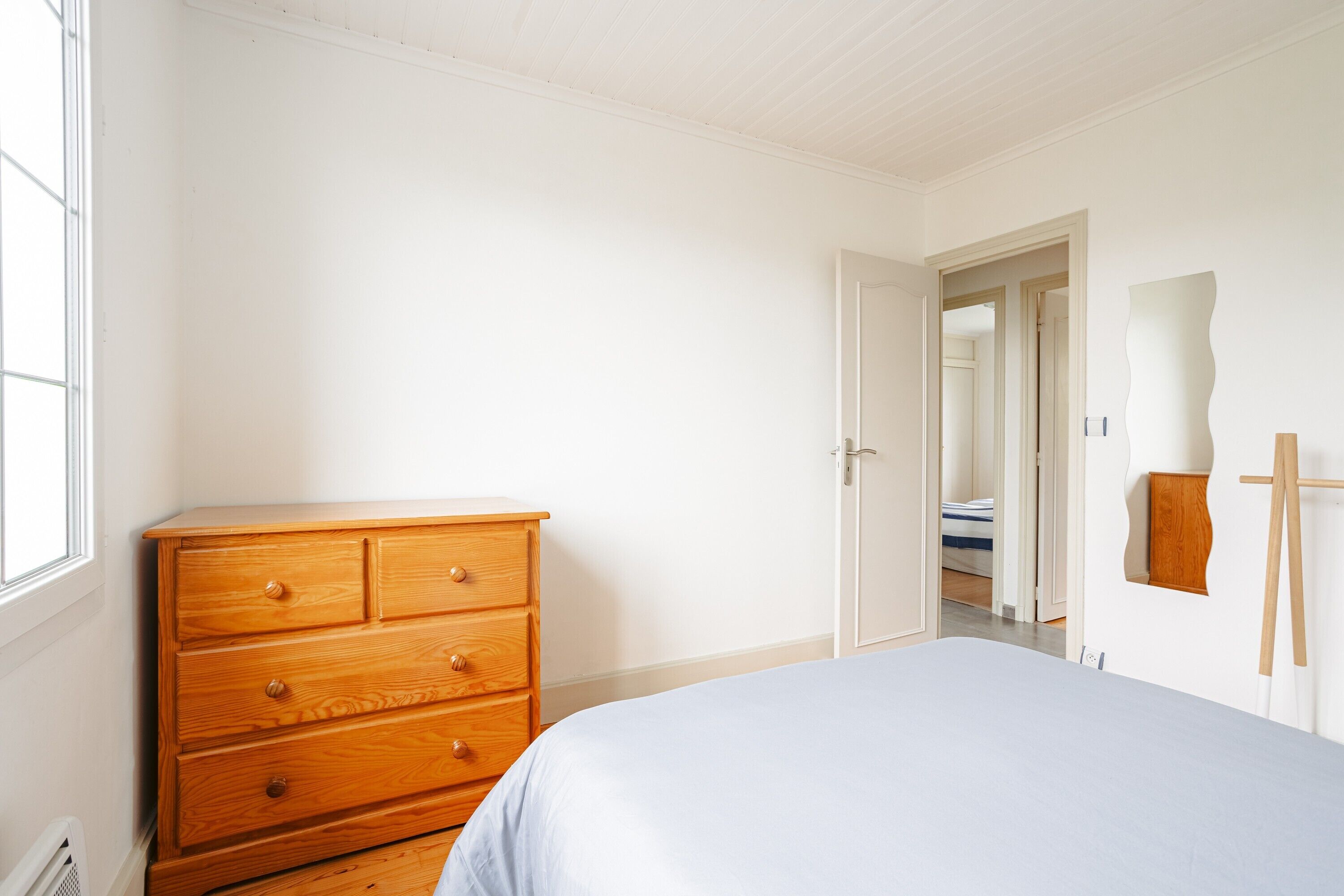Une chambre avec une commode en bois, un lit, une porte donnant sur une autre pièce et un chevalet fixé au mur.