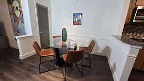 Un comedor con una mesa de vidrio y cuatro sillas marrones. Hay un cuadro en la pared y una zona de cocina con un salpicadero de azulejos.