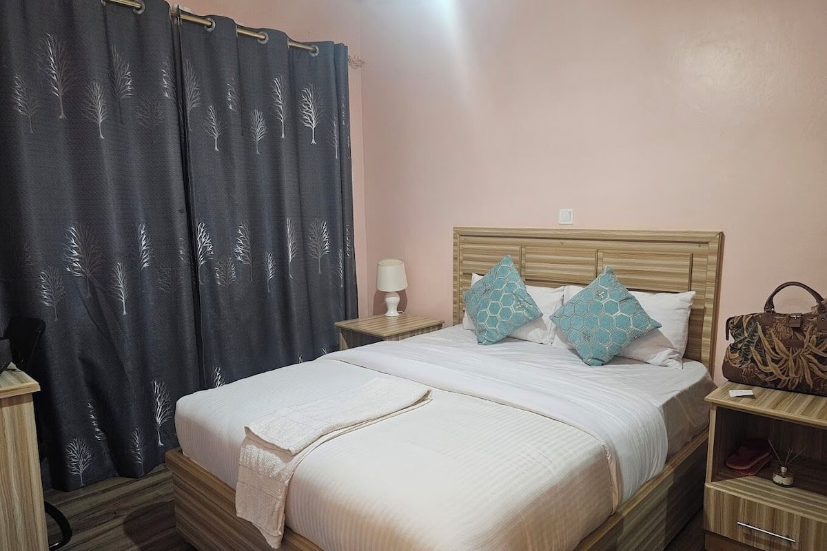 A bedroom with a bed, a nightstand, a lamp, a wooden headboard, and a curtain with a tree pattern.