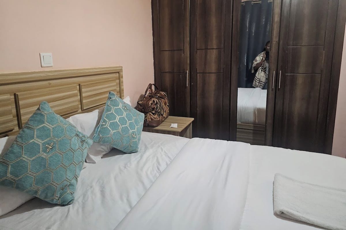 A neatly made bed with patterned pillows, a wooden headboard, a small bedside table with a lamp, and a dark wooden wardrobe.