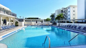 Una piscina con sillas de descanso y sombrillas, rodeada de edificios con balcones y palmeras.
