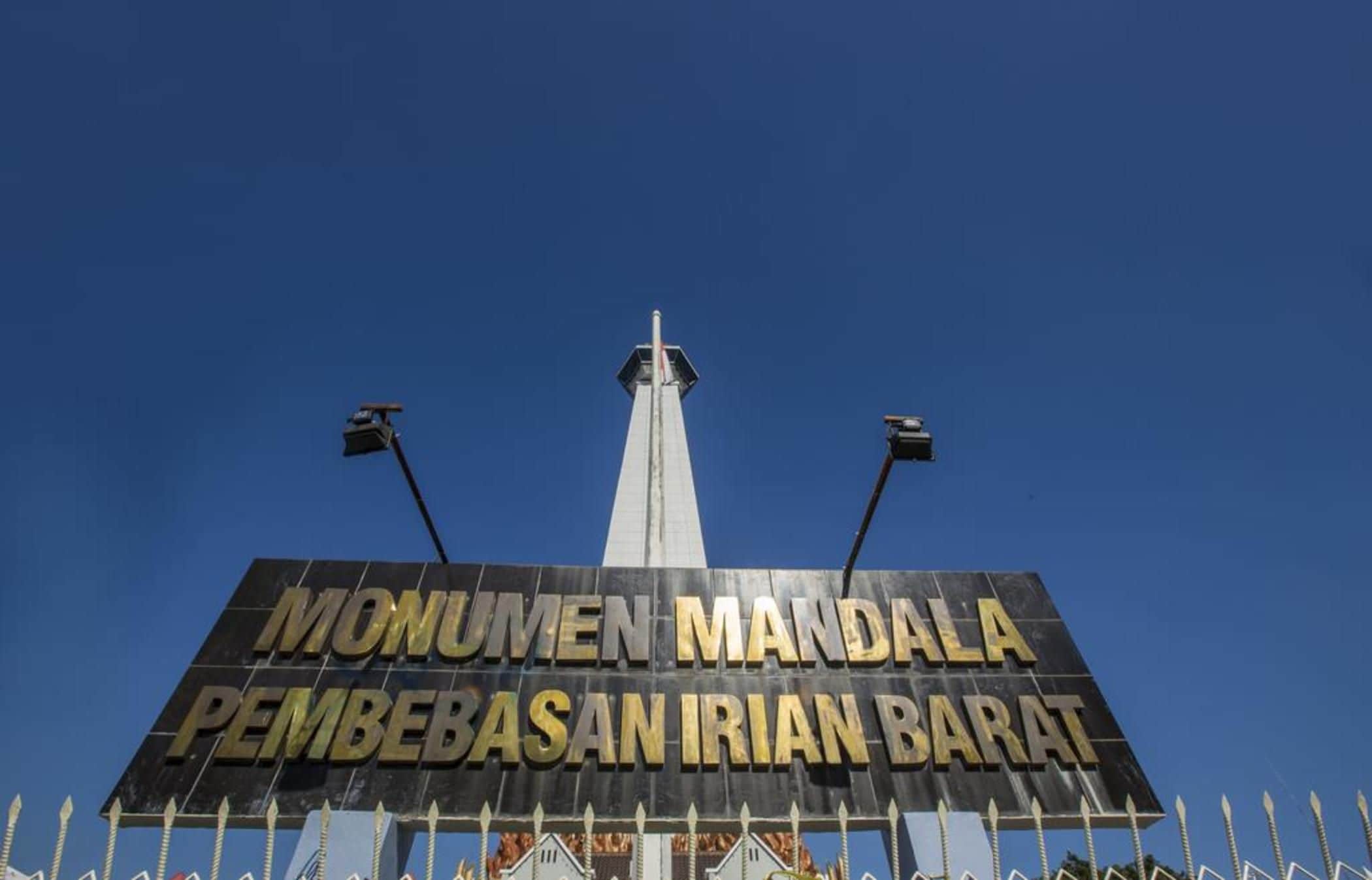 Een groot bord met de tekst 'MONUMEN MANDALA PEMBEBASAN IRIAN BARAT' tegen een helderblauwe lucht.