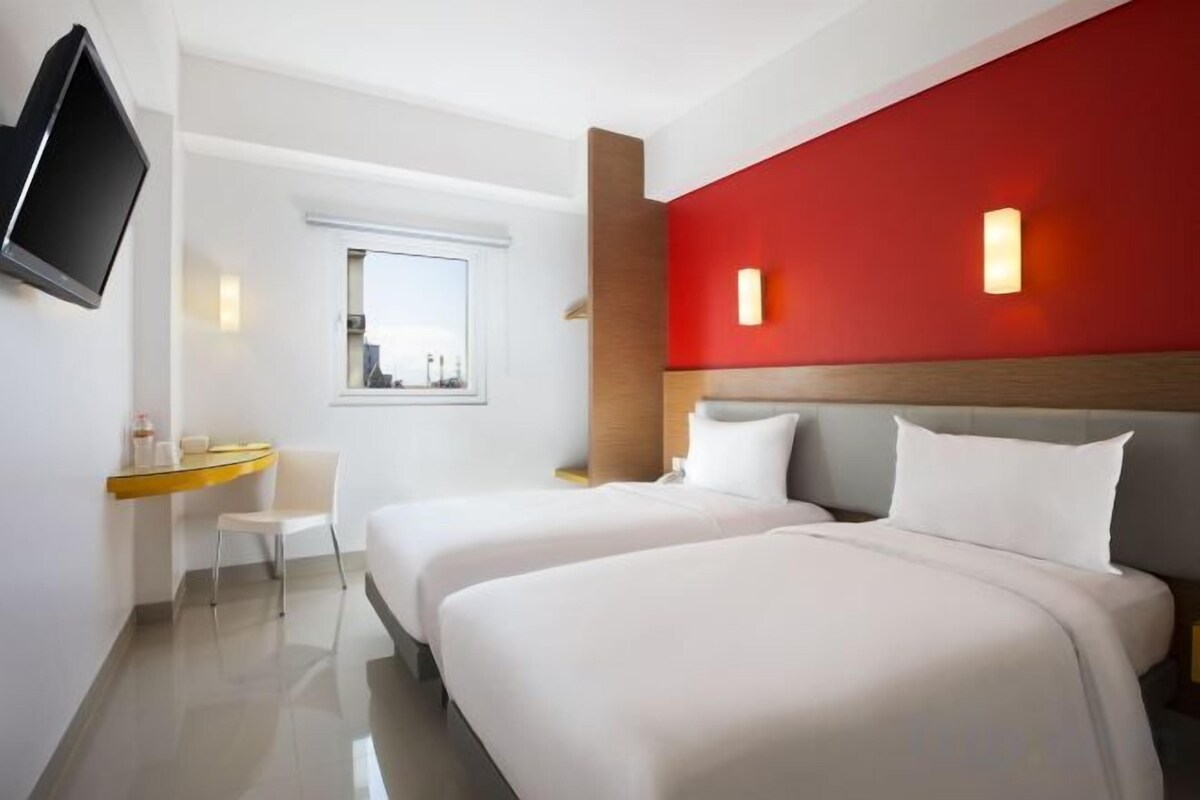 Une chambre d’hôtel avec deux lits, un bureau, une chaise, une télévision et un mur d’accent de couleur rouge.