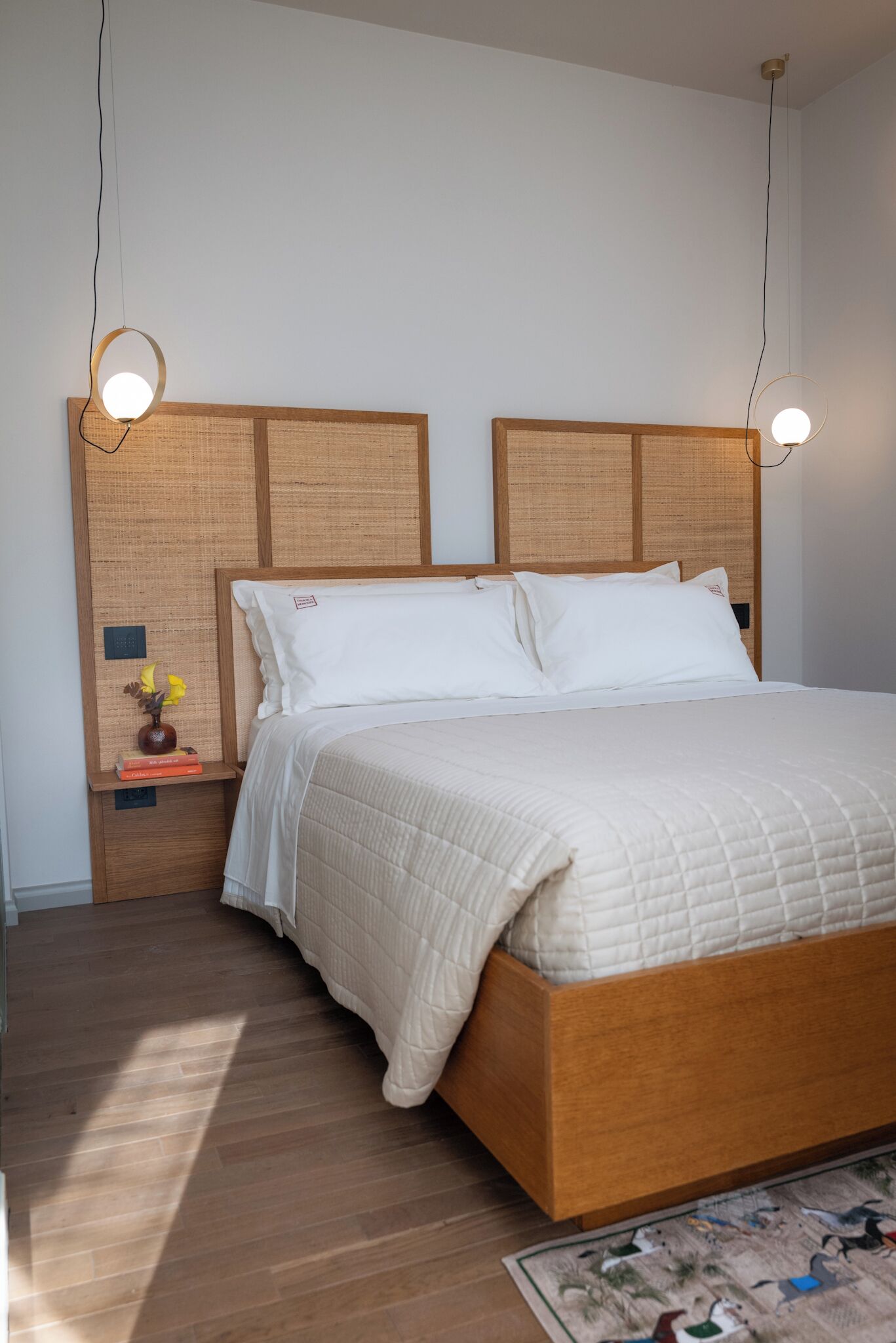 Ein ordentlich bezogenes Bett mit Holz-Kopfteil, ein Nachttisch mit Blumenvase und Büchern sowie zwei Pendelleuchten.