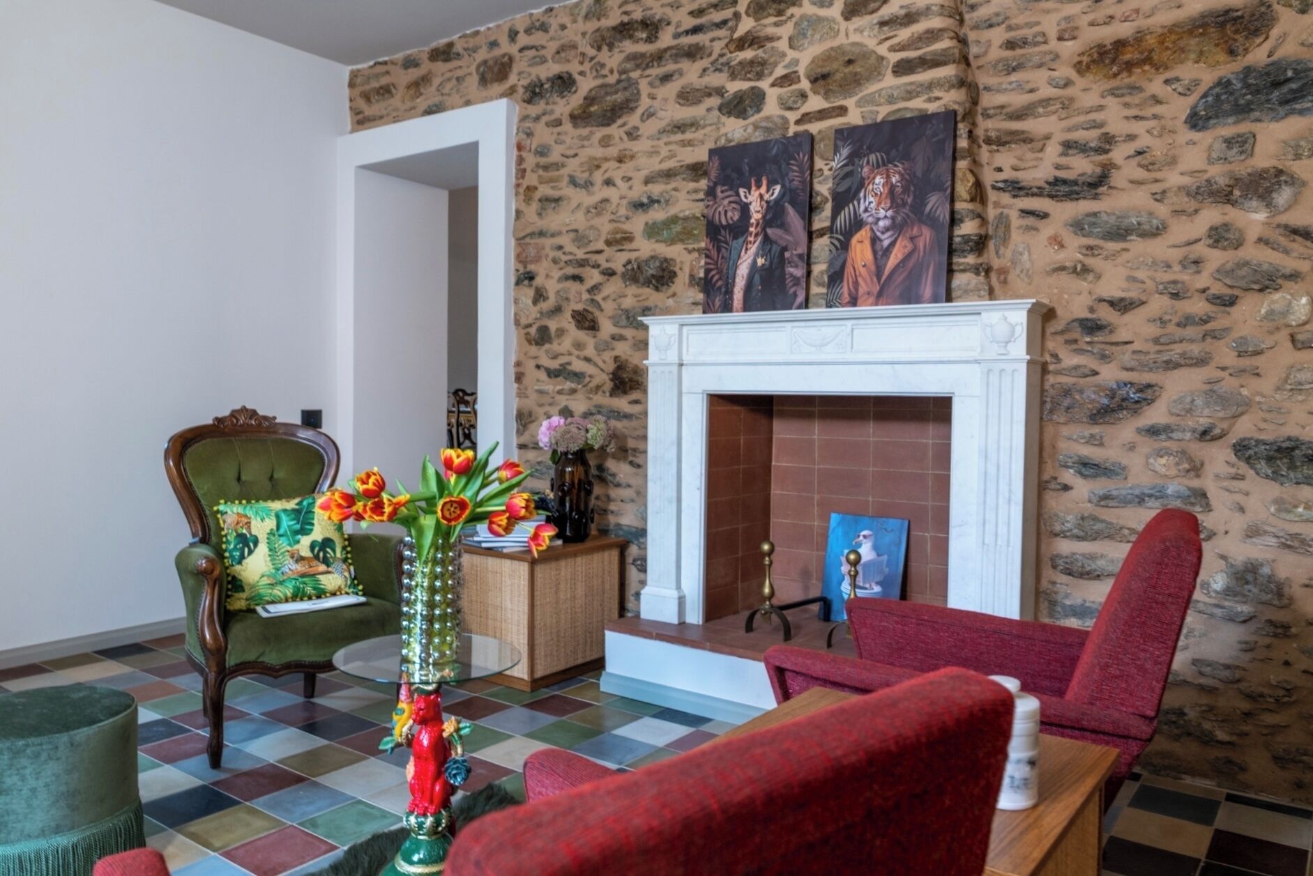 A room with a stone wall, a fireplace, a green armchair, a red armchair, and a vase with flowers.