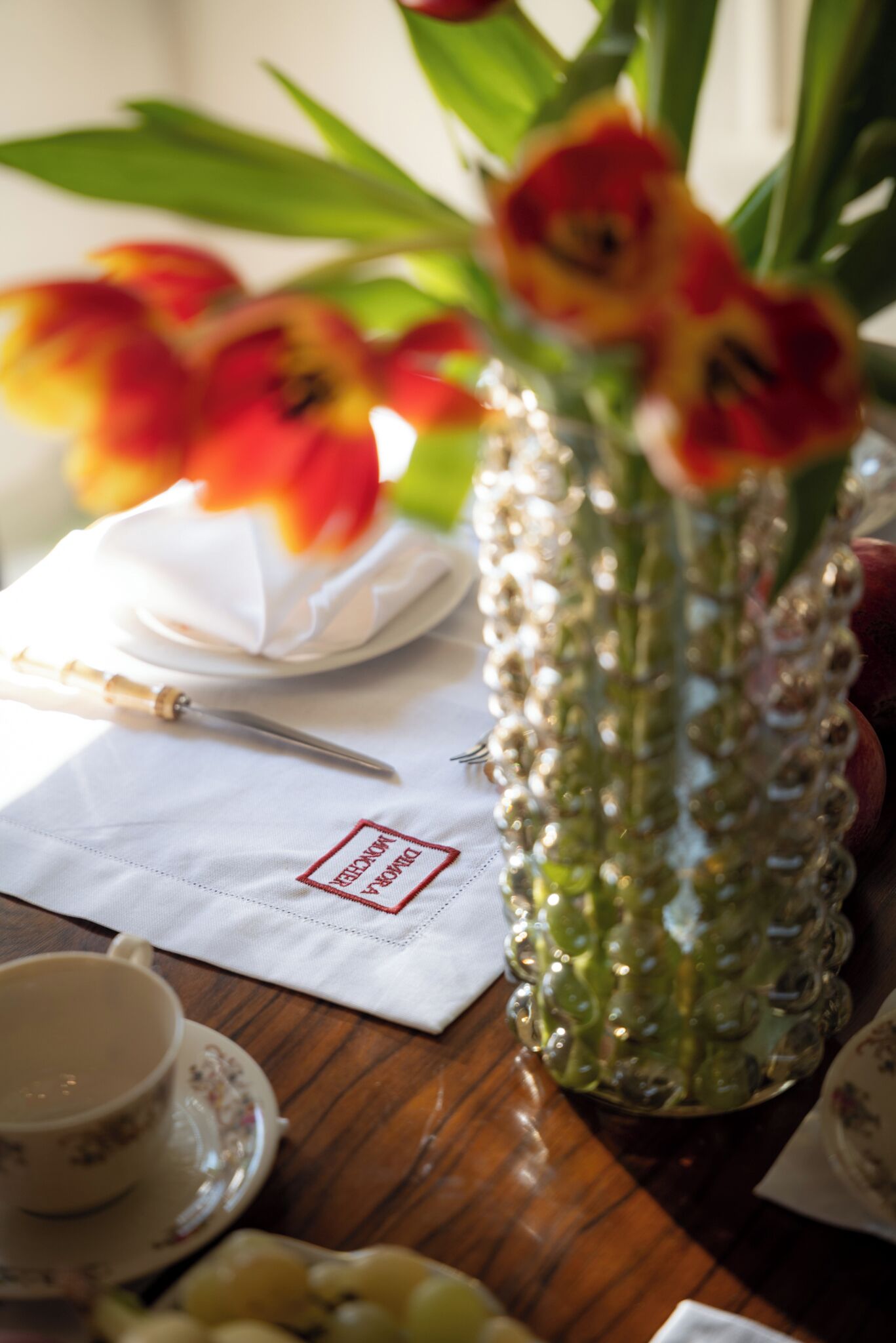 Una mesa con servilleta blanca, un florero de cristal con flores, una taza y un plato con fruta.