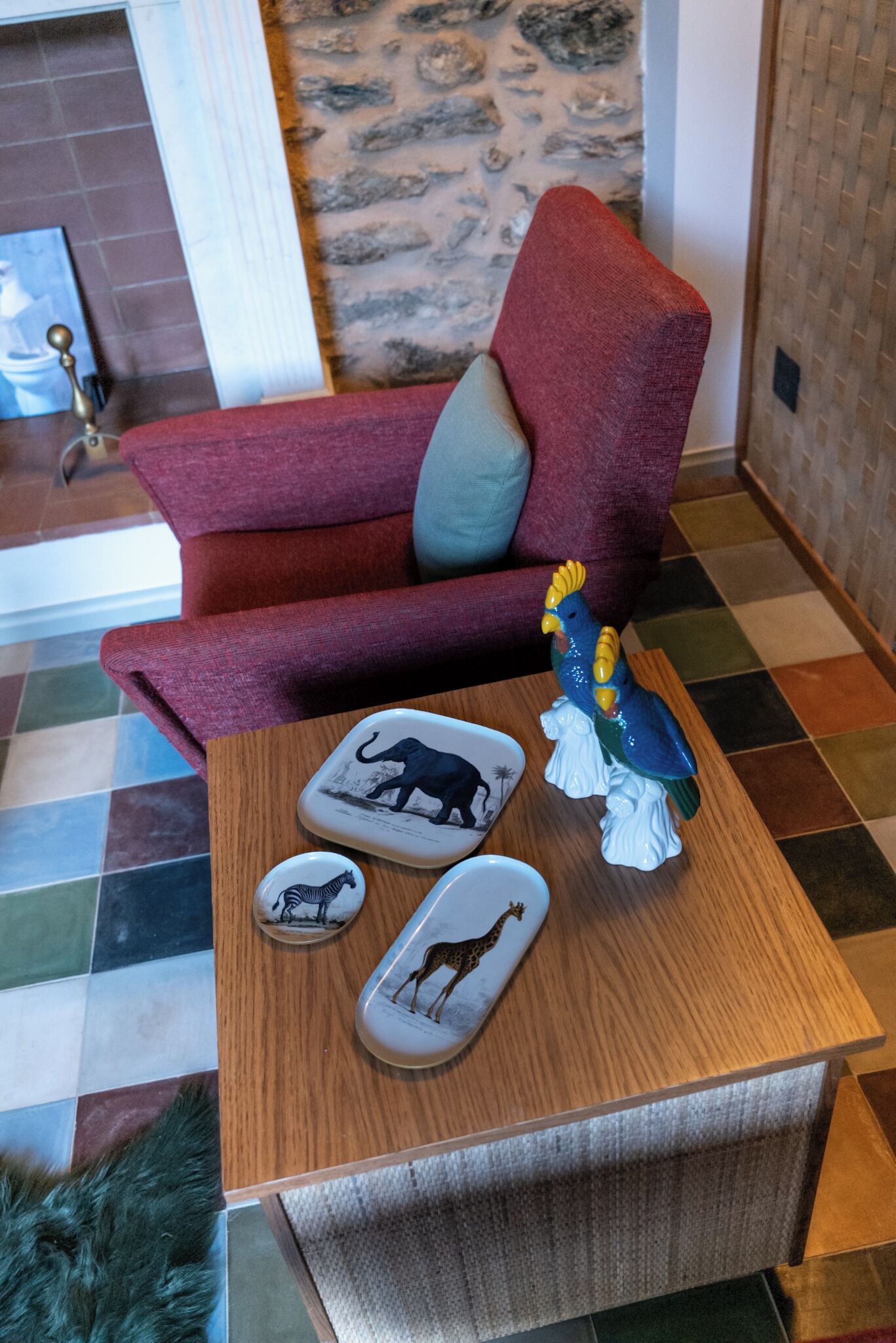 Un canapé rouge avec un coussin gris, une table basse en bois avec des dessous de verre en forme d’animaux et une statuette de coq décorative.