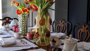 Een eettafel gedekt met witte tafelkleden, een vaas tulpen en divers servies, waaronder een theepot, kopjes en borden.