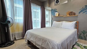 A bedroom with a bed, a standing fan, a window with curtains, and wall art. - Cozy Corner: Pet-Friendly 1BR with Sweeping City View (Taguig)