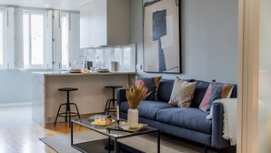Una sala moderna con un sofá azul oscuro, una mesa de centro y un jarrón con plantas secas. Hay una zona de cocina con una encimera blanca y taburetes de bar.
