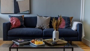 Una sala de estar moderna con un sofá azul oscuro, una mesa de centro con un libro, vasos y pasteles, y un florero con plantas secas.