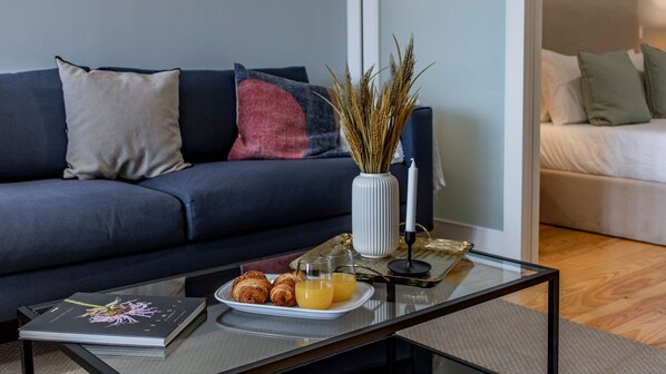 Een moderne woonkamer met een donkerblauwe bank, een glazen salontafel, een boek, croissants en sinaasappelsap.