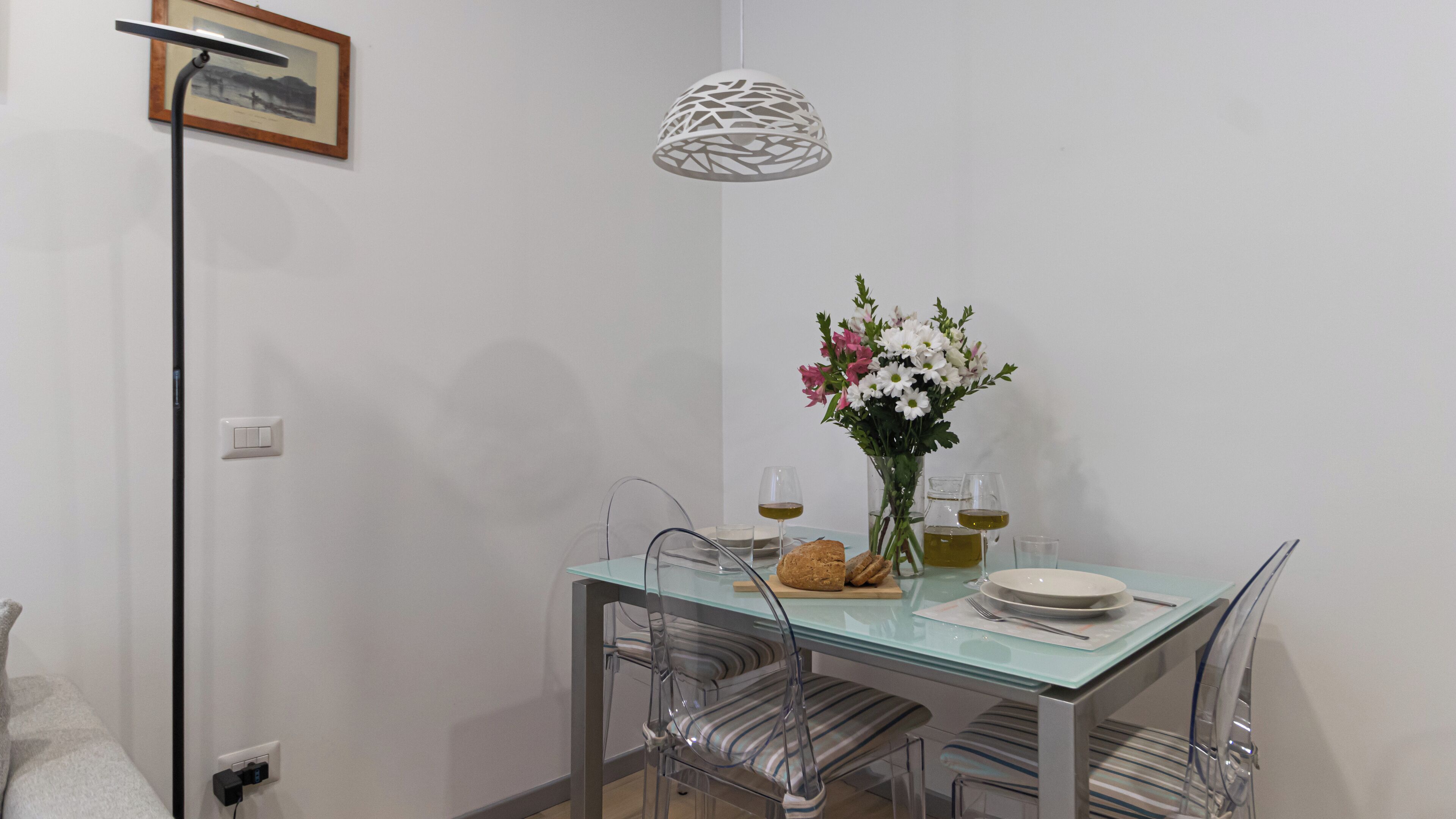 A dining area with a glass table, clear chairs, a white pendant light, and a vase with flowers.