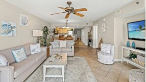A spacious living room with a ceiling fan, a television, and a kitchen in the background. - Crescent Shores 309- Oceanfront 2 BBR- Pool (North Myrtle Beach)