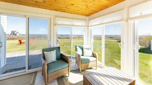 A covered porch with wicker furniture, large sliding glass doors, and a view of the ocean. - Shore to Sky Oceanfront Escape with Sunroom, Stunning Views & Beach Access (Prince Edward Island)