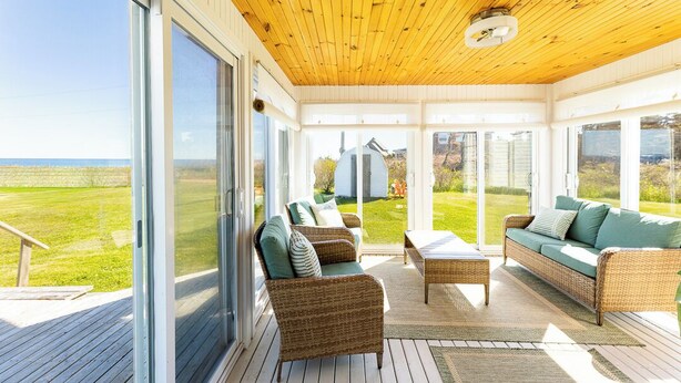A covered outdoor deck with wicker furniture, large windows, and a view of the landscape. - Shore to Sky Oceanfront Escape with Sunroom, Stunning Views & Beach Access (Prince Edward Island)