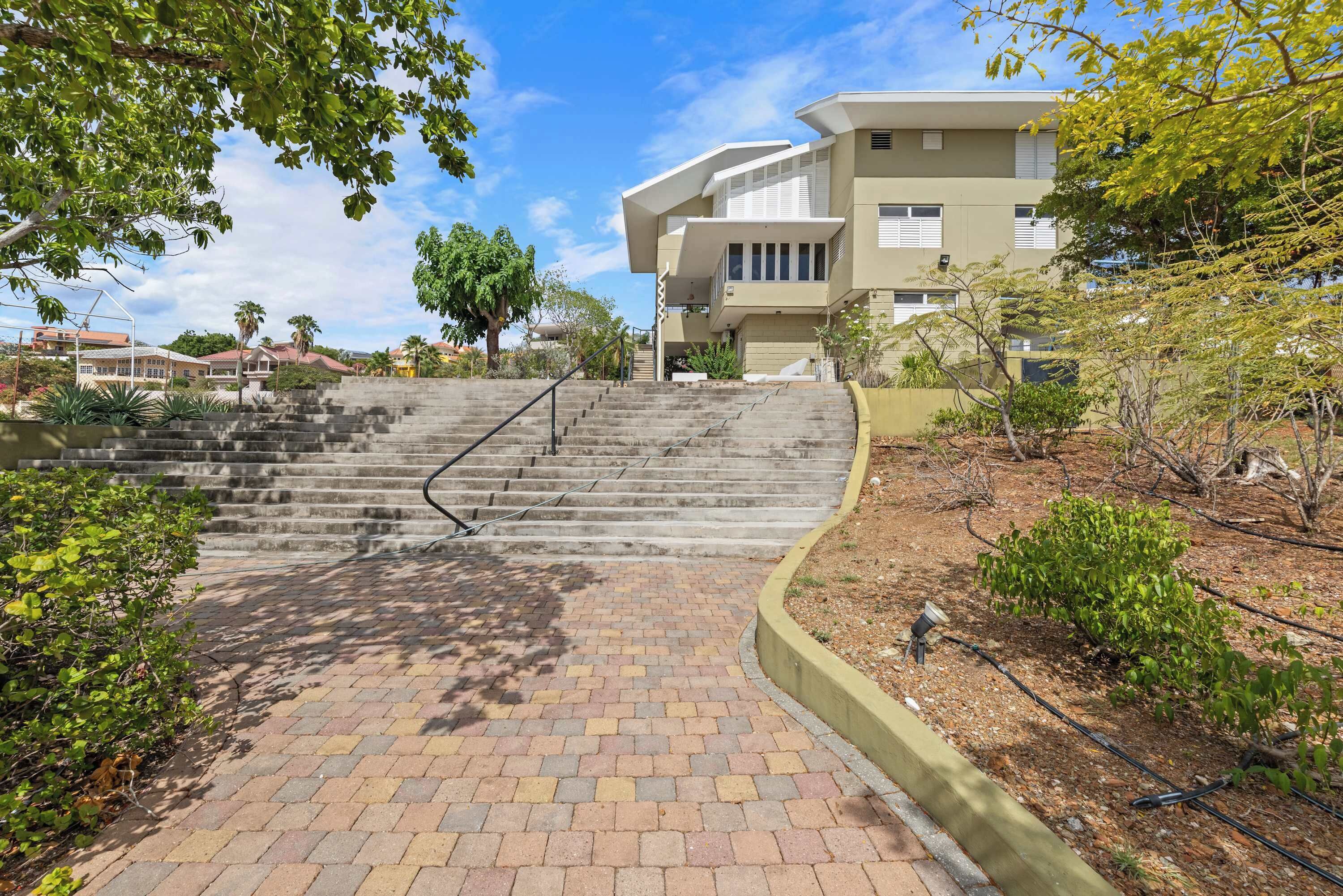 A modern building with a staircase leading to the entrance, surrounded by greenery and a paved pathway.