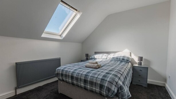 A bedroom with a sloped ceiling, a skylight, a bed with a plaid duvet, a bedside table with a lamp, and a radiator.