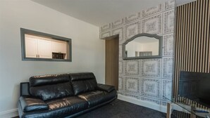 A room with a black leather sofa, a television on a wooden stand, and a patterned wall with a mirror.