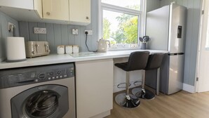A compact kitchen with a washing machine, a toaster, and a refrigerator. There are two bar stools and a window providing natural light. - Pet Haven Humberston Fitties (Humberston Fitties Cleethorpes)