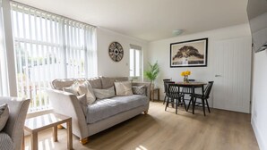 A living room with a sofa, a dining table with chairs, a clock on the wall, and a door. - Pet Haven Humberston Fitties (Humberston Fitties Cleethorpes)