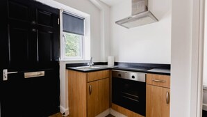 A compact kitchen with a black door, a window with a white blind, a stainless steel range hood, and wooden cabinets. - Withernsea Studio Apt + Sofa bed (Withernsea)