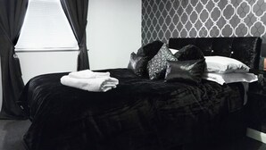 Ein ordentlich bezogenes Bett mit schwarzer Bettdecke und Kissen, ein Nachttisch mit Lampe sowie eine Wand mit Muster im Hintergrund.