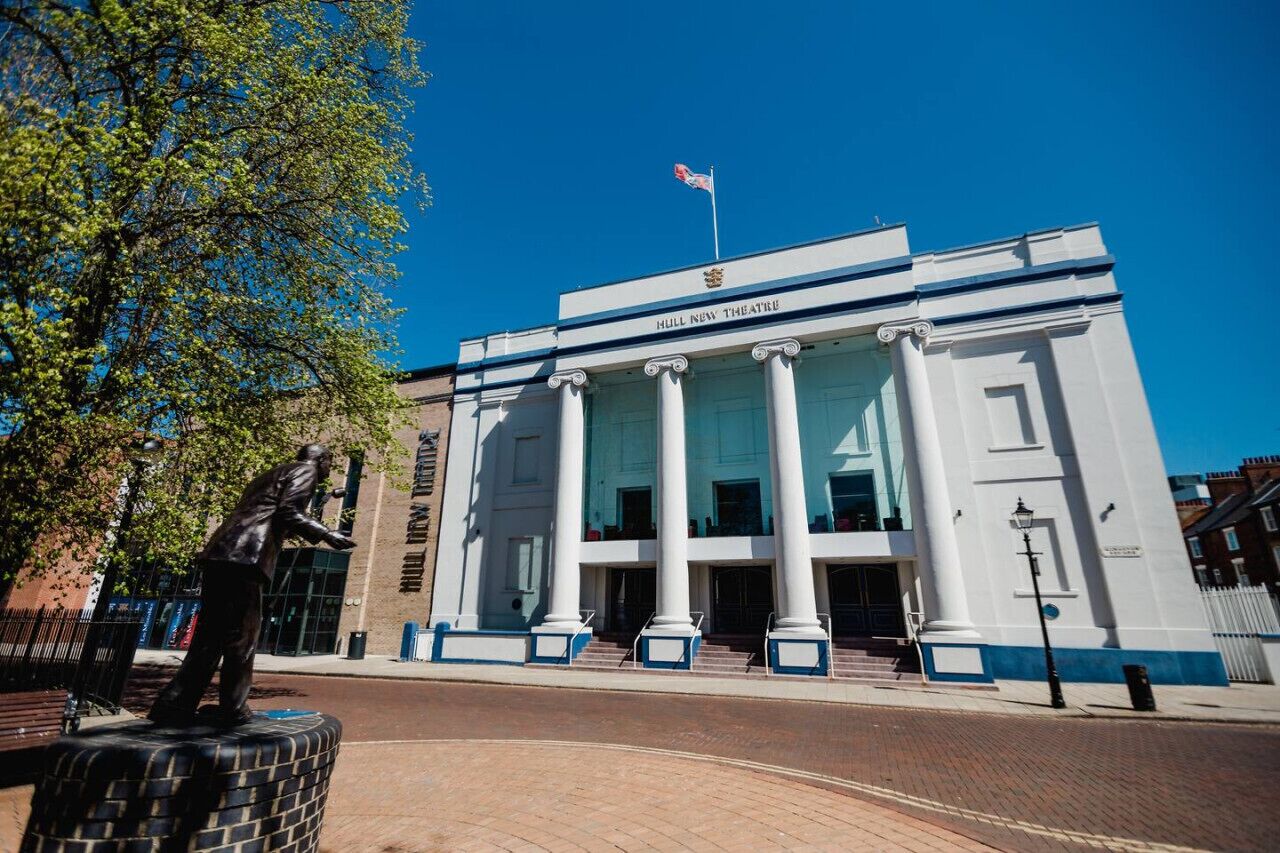 The exterior of the Hull New Theatre with classical columns, a statue, and a clear blue sky.