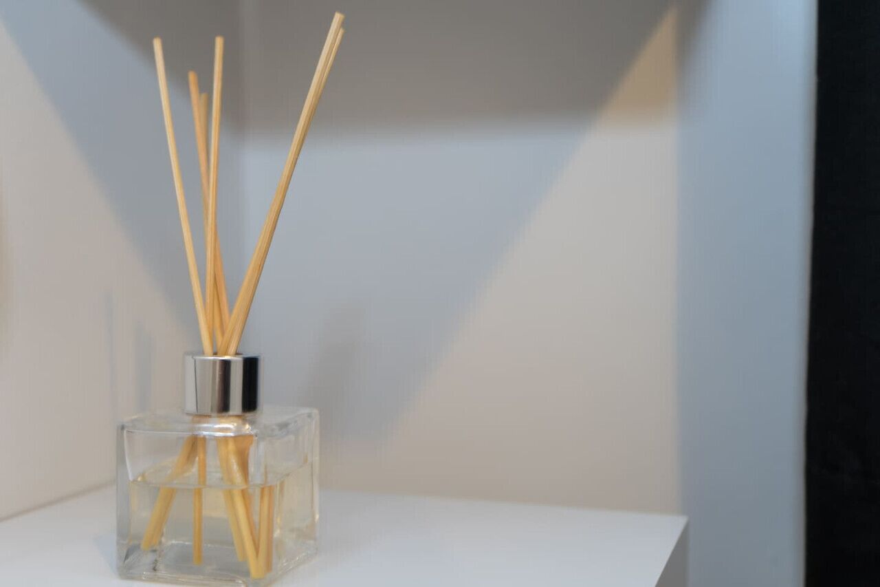 Eine klare Glasflasche mit Holzstöckchen auf einer weißen Oberfläche vor neutralem Hintergrund.