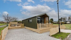A small, single-story wooden cabin with a sloped roof, a wooden deck, and a gravel driveway. - Lowfield Lodge & Hot Tub (Humberston Fitties Cleethorpes)