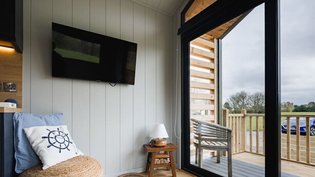 A cozy room with a TV mounted on the wall, a wooden bench, a small table with a lamp, and a large woven ottoman. - Lowfield Lodge & Hot Tub (Humberston Fitties Cleethorpes)
