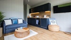 A modern kitchen with dark cabinets, a flat-screen TV, and a dining area with a blue sofa and wicker ottoman. - Lowfield Lodge & Hot Tub (Humberston Fitties Cleethorpes)