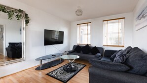 A modern living room with a flat-screen TV, a glass coffee table, a grey sofa, and a large mirror with greenery. - Hullidays》Victoria Dock Residence (Kingston upon Hull)