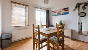 A dining area with a wooden table and chairs, a radiator, a window with blinds, a painting on the wall, and a clock. - Hullidays》Victoria Dock Residence (Kingston upon Hull)