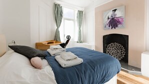 Un dormitorio con cama, mesita de noche, sillón, chimenea y un cuadro en la pared.