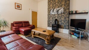 A living room with a red leather sofa, a wooden coffee table, a TV stand with a flat-screen TV, a bookshelf, and a wall-mounted fireplace.